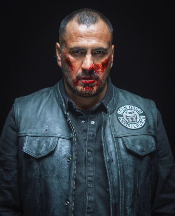 Tammy Davis wears a black leather jacket and looks grimly at the camera with blood smeared on his face.