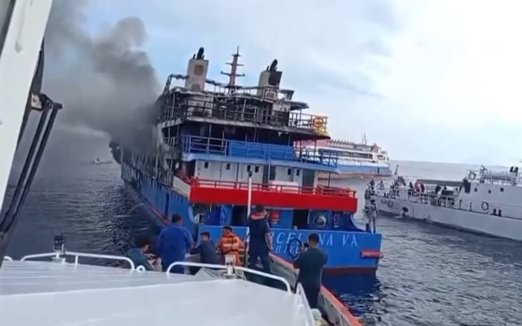 Dozens of people can be seen floating in the ocean, watching the KM Barcelona 5 ferry as it burns.