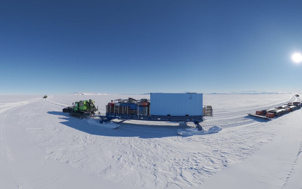 A lime-green heavy vehicle on caterpillar tracks makes its way across a snowy plain in Antartica, towing a large sled laden with freight containers.