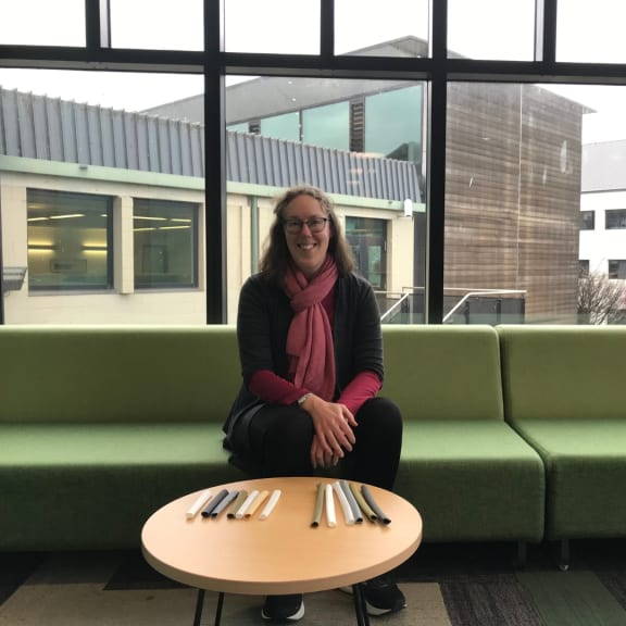 A woman wearing a red scarf sits on a green couch smiling at the camera. Behind her are large windows looking out to more buildings. In front of her is a round coffee table with 3D-printed flutes of various sizes and colours on it.