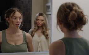 Two young blonde women stand in front of a bathroom mirror while giving each other a significant look.