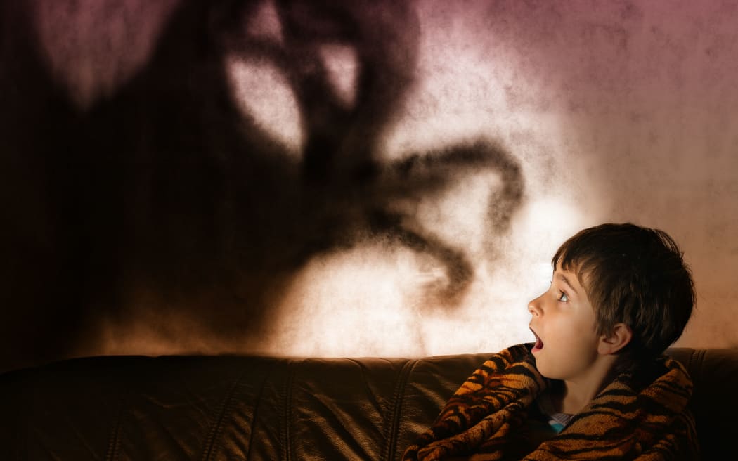 A photo of a boy in the dark with a shadow behind him in the shape of a monster with its arms extended.