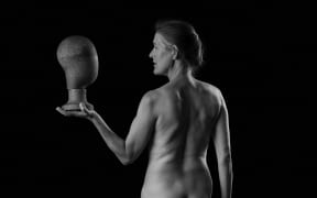 A woman, over 50, stands with her bare back to the camera. She's looking to the left side at a smooth mannequin head held in one hand.