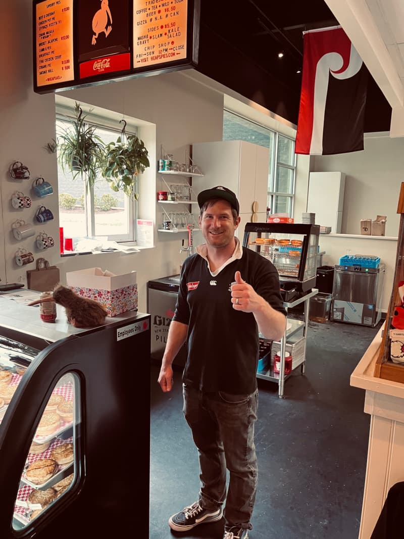 A man dressed in a black rugby jersey and cap stands in the kitchen of a pie shop. The tino rangatiratanga flag is hung up in the background.