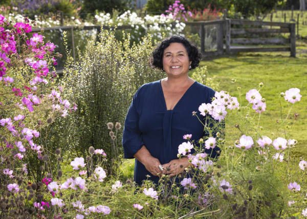 Pounamu Skelton stands smiling in a beautiful garden amongst colourful flowers.