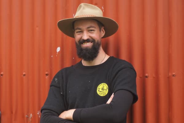A man wearing a black long-sleeved shirt and a tan-coloured wide-brimmed hat stands with his arms folded in front of burnt orange corrugated iron. He has a dark beard and is smiling warmly.
