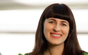 Jane sits on a chair and smiles at the camera. She wears a vibrant green top and a large green ring.