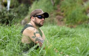 Josh McNally sitting in long grass, wearing sunglasses and a cap.