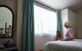 An elderly woman sitting on her bed looking out the window