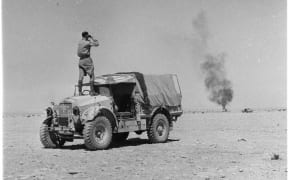 Mobile Unit commentator Arch Curry standing on a jeep at Alamein, viewing a burning New Zealand jeep through binoculars