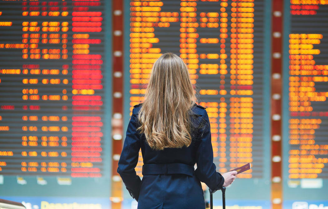 Young woman in international airport looking at the flight information board, holding passport in her hand, checking her flight