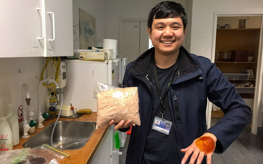 A man standing next to a kitchen sink with a fridge in the background. The man is smiling and holds a plastic bag of a coarse off-white substance and has a circular piece of orange "skin" on his outstretched hand.