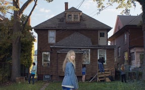 A young woman stands in front of a shadowy and dilapidated brick house.