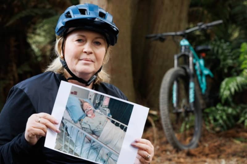 Rotorua woman Kath Cross, in her mountain biking gear in a forest, holds up a printed picture of herself from when she had lung transplant surgery.