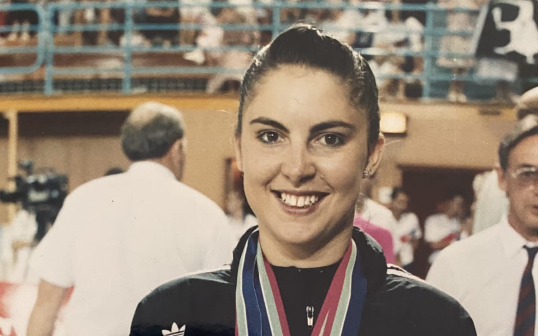 Angela Walker is wearing her NZ Team jacket and standing in the gymnastics arena at the 1990 Commonwealth Games. She is smiling at the camera and wearing three of her medals around her neck.
