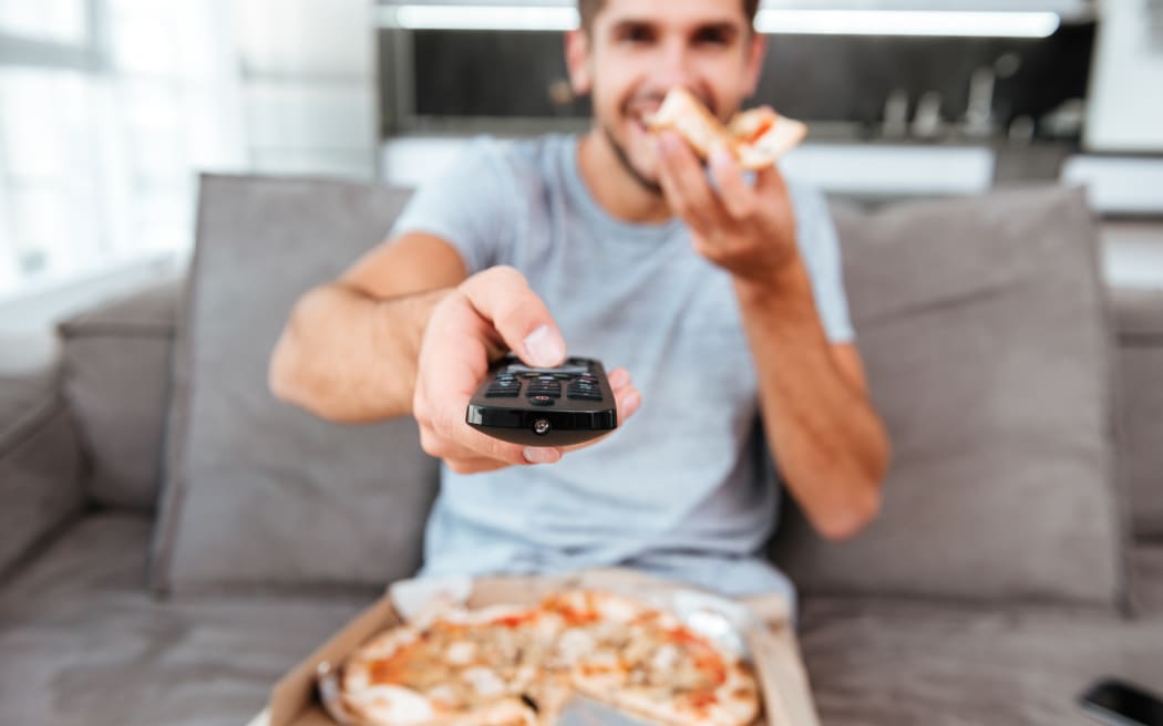 Image of young joyful man holding remote control and pushing the button while eating pizza. Focus on remote control.