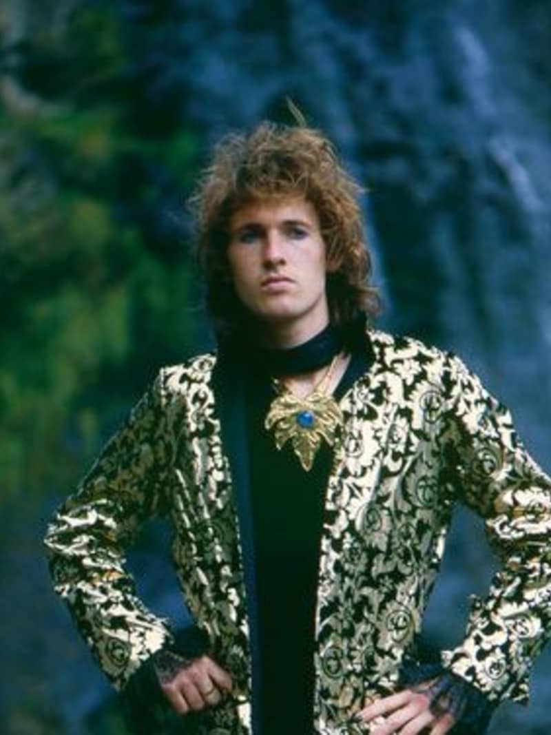 An image of New Zealand musician Andrew Fagan pictured in front of a waterfall. Andrew has shaggy brown hair and is wearing a shiny black and gold suit, with a black shirt and elaborate gold corsage.