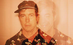 American musician Bill Callahan wearing a cap and patterned shirt in an image with an orange filter