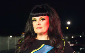 A woman with long black hair wearing a black top with a colourful lightning bolt gazes at the camera.