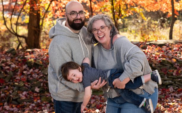 Picture shows a family portrait against golden leaves as a background. A man and a woman smiling at the camera, with their son between their arms.