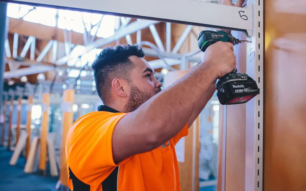 A man in a fluorescent orange shirt drills a nail into a structure. He is standing in a construction site.