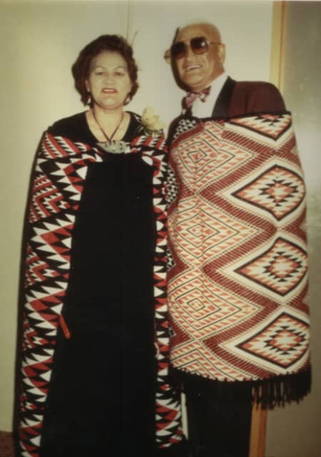 A couple wear woven cloaks and smile together against a white wall.