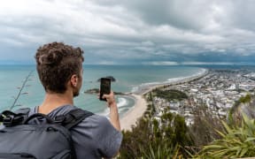 A man takes a photo of Mount Maunganui beach in the Bay of Plenty with a smartphone.