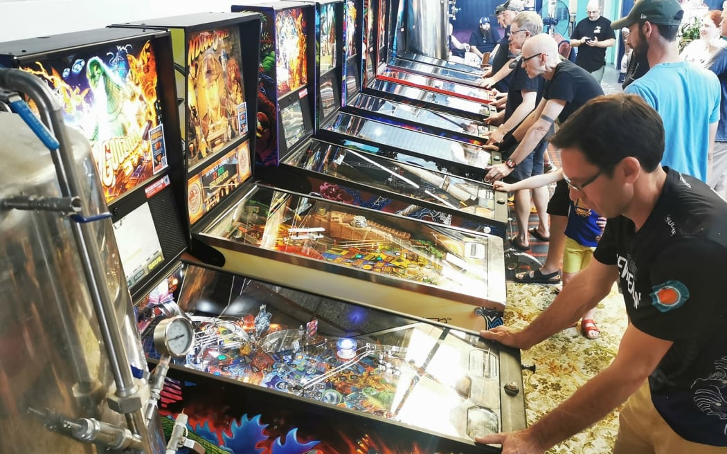 This image shows a line of pinball machines along the left hand wall. All the machines are being played, and there is a crowd of people milling around behind them.