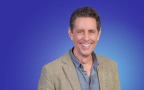 Host Wallace Chapman in a blue shirt and casual jacket. Soft blue background.