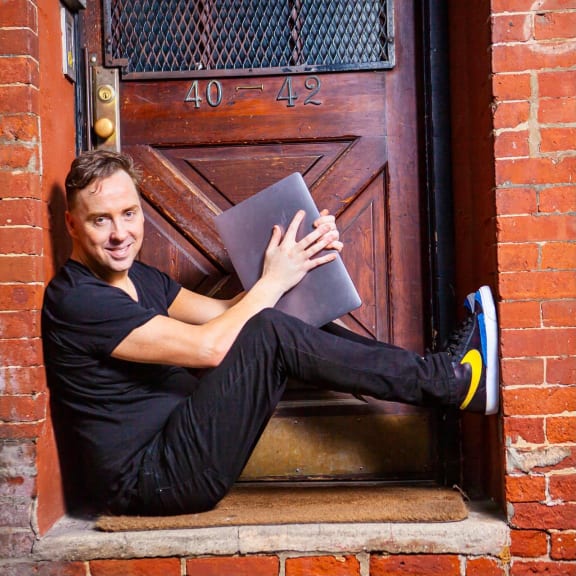 Geoffrey John Davies, founder of The Violin Channel, sits on the ground sideways across a doorway. He's holding a laptop computer.