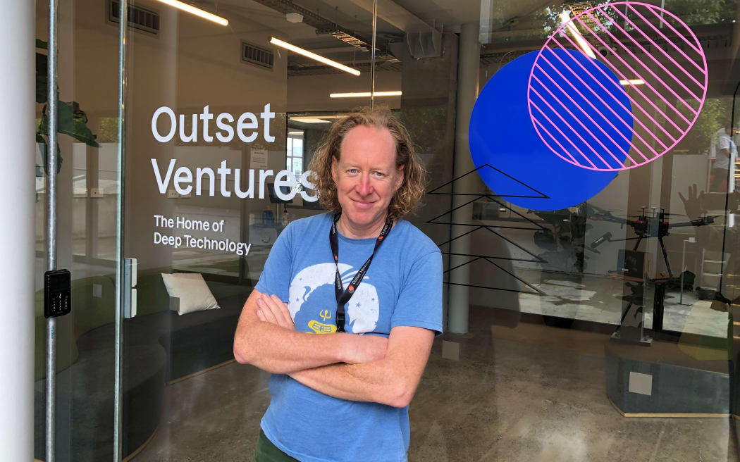 Sean smiles at the camera. He is standing outside an office building. The window of the business says "OUTSET VENTURES - THE HOME OF DEEP TECHNOLOGY".