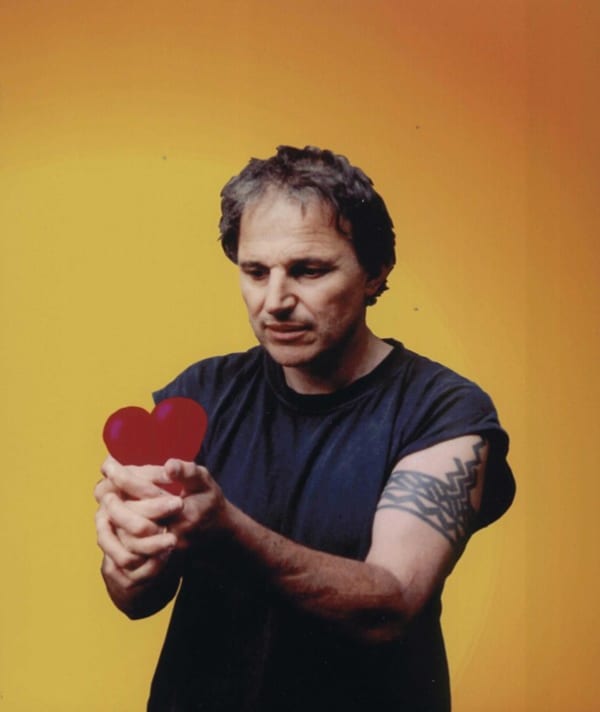 Chris Knox, wearing a black t-shirt, holds a red paper heart and gazes at it.