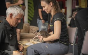 A woman cradles a kiwi on her lap while a man looks at the kiwi.