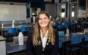 A teenage girl with long brown hair wearing a school uniform blazer with badges on the lapels, and a pounamu necklace, sits in a classroom smiling at the camera.