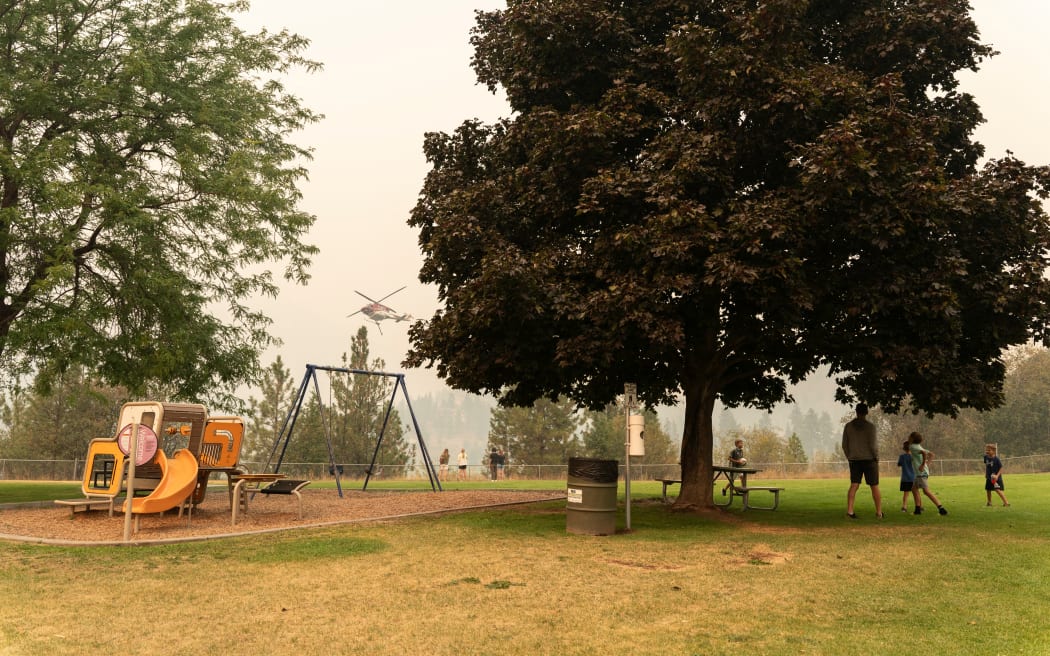 People gather at a lookout as a helicopter gathers water from Shannon Lake to fight the McDougall Creek wildfires in West Kelowna, British Columbia on 19 August, 2023. Residents of western Canada scrambled to evacuate as raging wildfires encroached on two metropolitan areas - separate blazes that have sent tens of thousands fleeing over the course of just days.