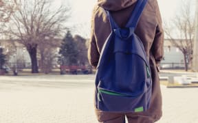 A photo of a teenager from behind showing her back pack and a school yard in the back ground