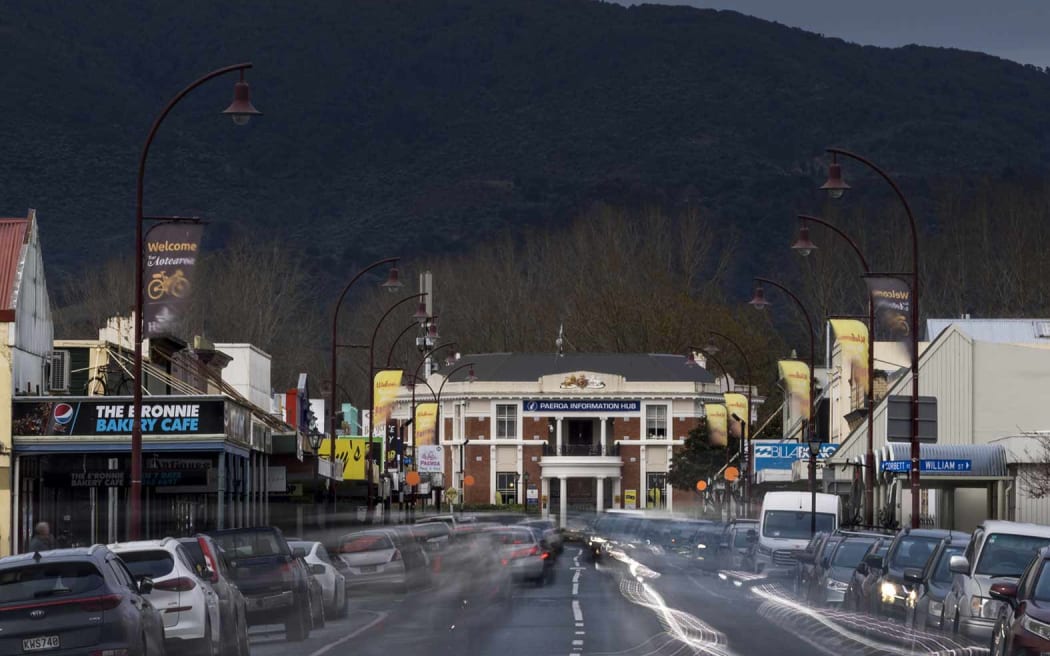 The main street of Paeroa below a grey sky. The photo has long-exposure, showing car headlights moving down the street in a ghostly way.
