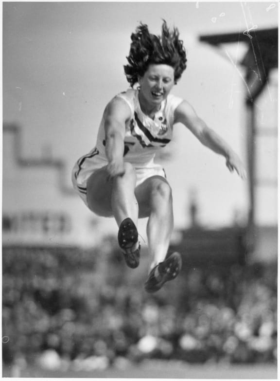 Yvette Williams in mid-air during a long jump at Carisbrook Park, Dunedin, in front of Queen Elizabeth II and Philip, Duke of Edinburgh, 26 January 1954.