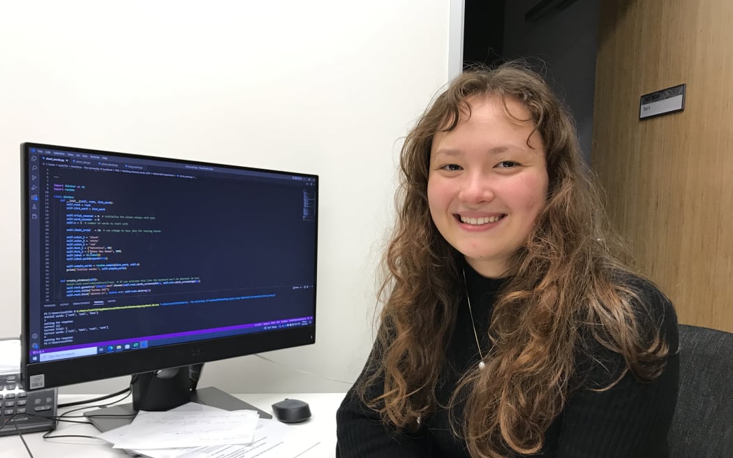 A woman with long curly brown hair sits at a desktop computer. She is turned towards the camera and smiling.