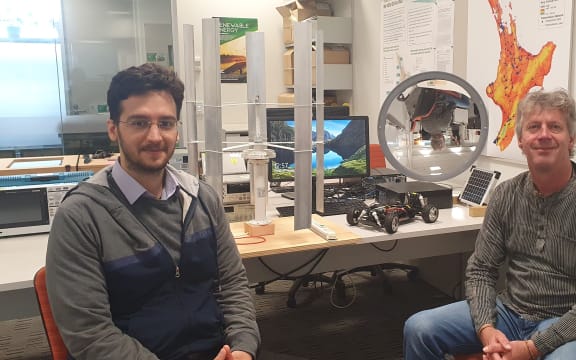 Soheil Mohseni and Alan Brent work with sustainable energy systems. There are mini versions of a vertical wind turbine and a solar concentrator on the desk behind them.