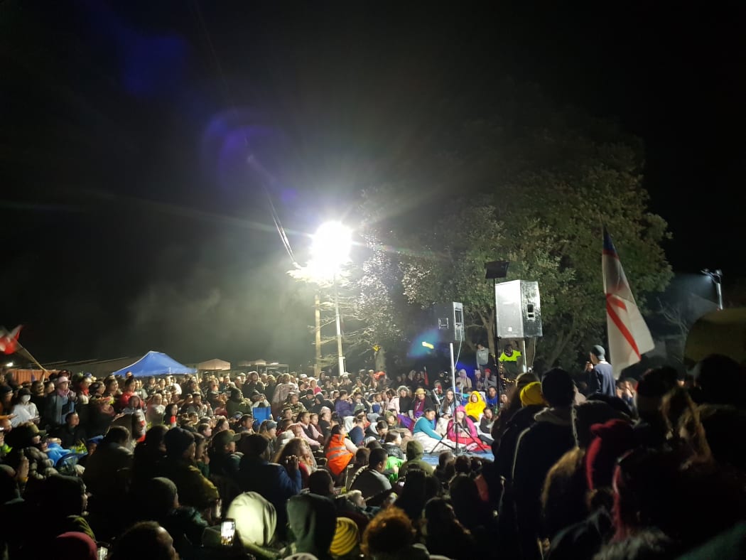 Thousands of people huddle around a small stage at Ihumātao enjoying a free concert.
