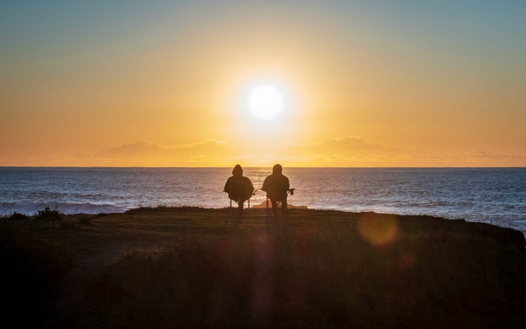A couple sit together on fold out chairs watching a sunset over the sea.