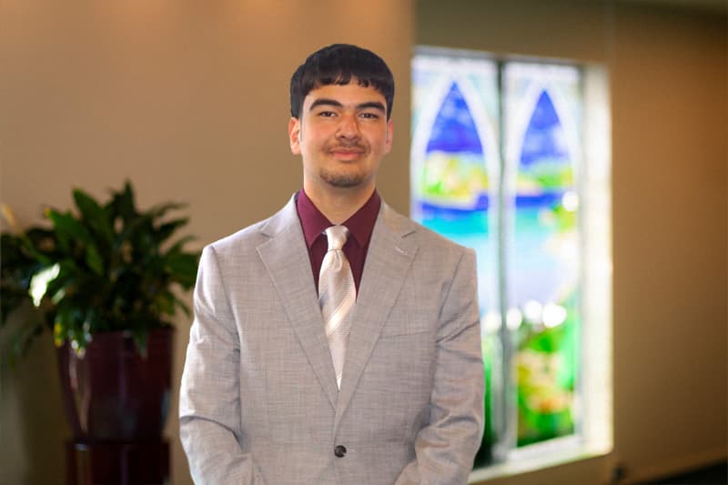 Harbour City Funeral Home assistant James Madden wearing a suit in a room with a stained glass window in the back.