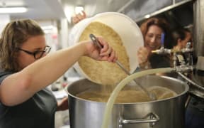 Two women pour grains from a bucket into a stainless steel tub.