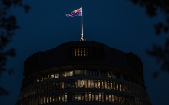 A night view of the Beehive, with interior lights on. The New Zealand flag is flying atop the building against a dark blue sky, framed by silhouetted tree branches.