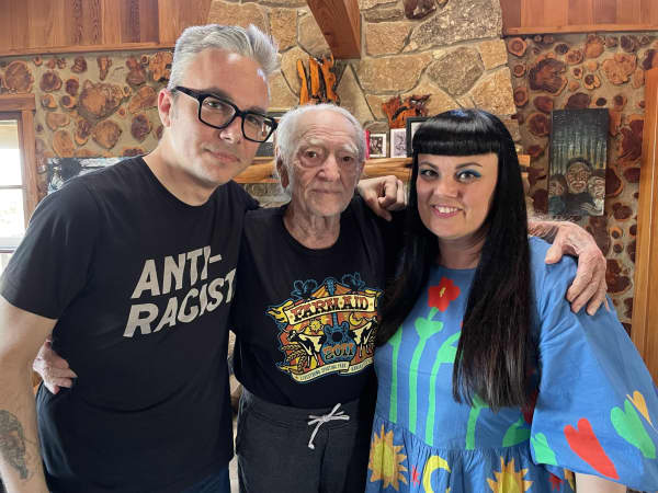 Two white haired men and a black haired women stand close together.