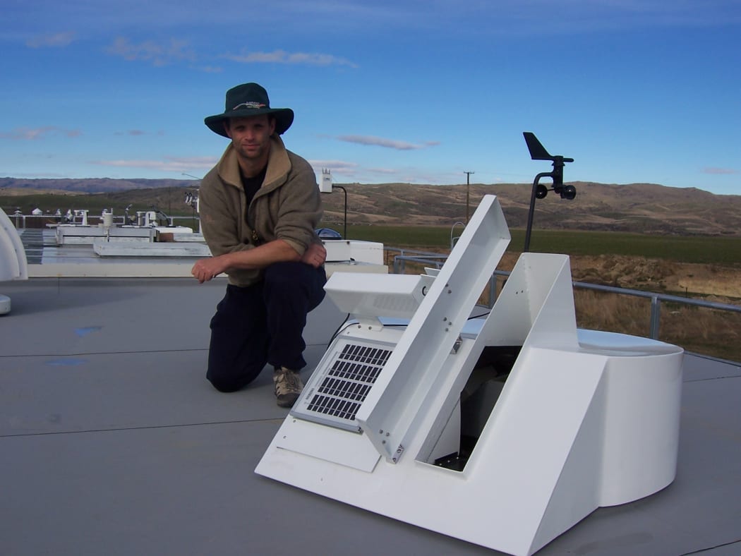 Dan Smale on top of the NIWA's Lauder atmospheric research laboratory building kneeling behind a solar tracker that feeds light into a Fourier transform spectrometer used to measure atmospheric trace gases.