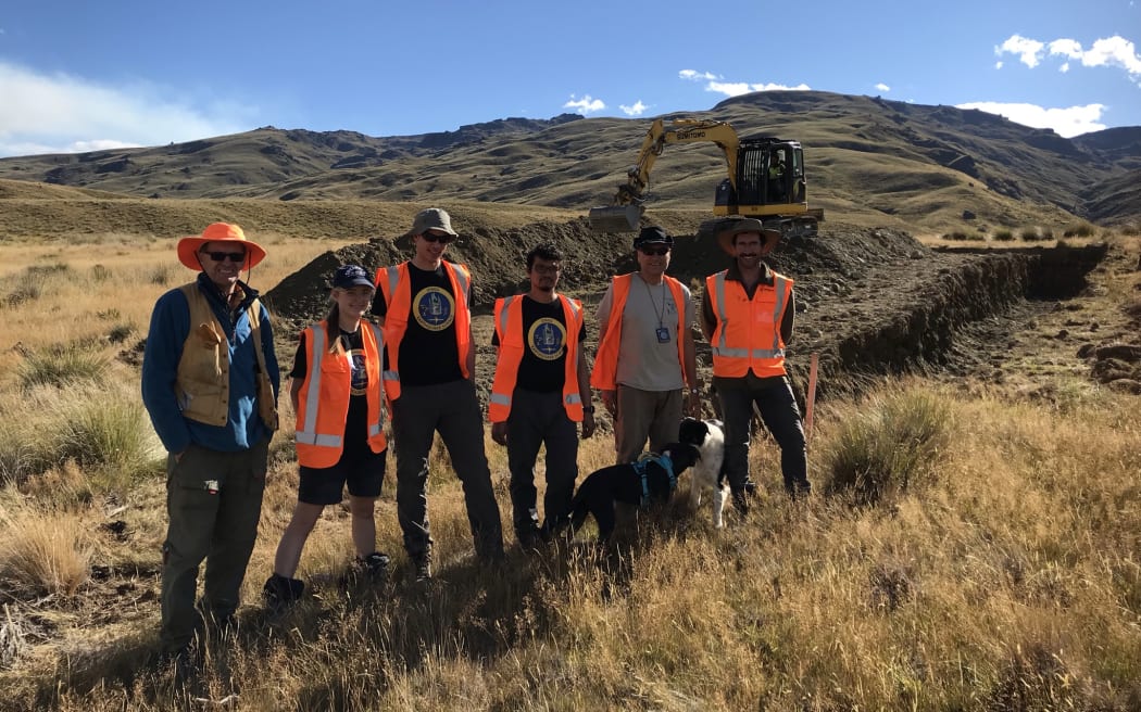The team of geologists and two dogs stand in front of a yellow digger which is digging a large trench in a grassy hilly landscape.