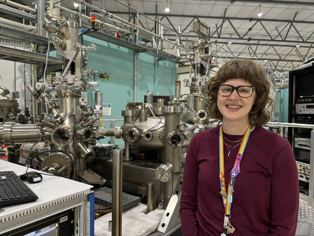 A woman with glasses and a rainbow lanyard standing in front of complicated metal machinery.