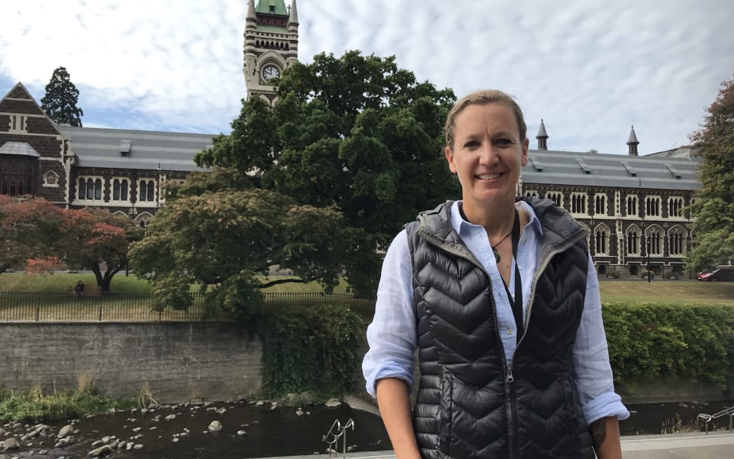 Janelle is standing in front of the Leith river with the University of Otago clocktower building in the background. She is smiling and wearing a puffer vest over a blue button up shirt.